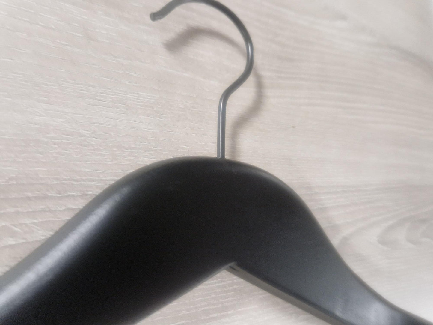 Close-up image of a stylish Hangers of London Wooden Jacket Black With Clips (Black Hook) 39cm hanging on a light-colored wooden surface. The metal hook of the hanger is prominently visible, curving over the edge of the surface. The wooden texture of the background is clearly detailed.