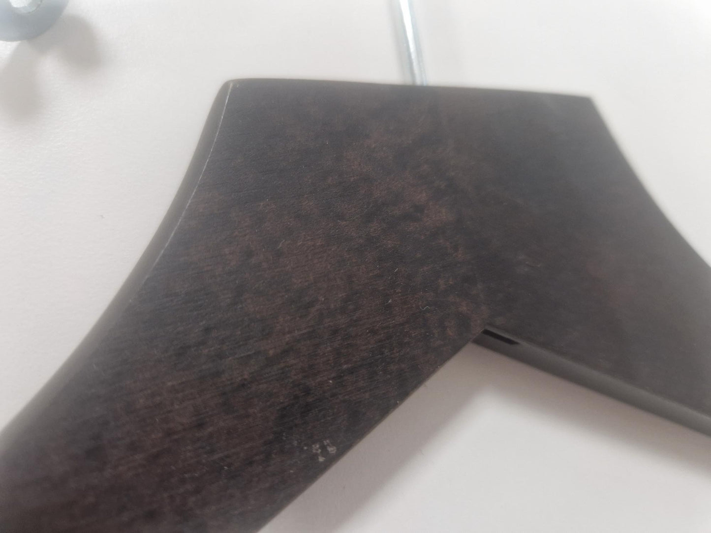 Close-up of the top part of a Hangers of London Wooden Hotel Anti Theft Jacket (Dark Brown) 44cm with a dark brown finish and metal hook, hanging against a light-colored wall. The hanger has a slightly curved, wide shoulder design.