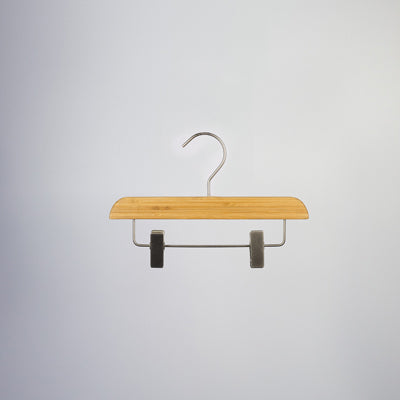A Hangers of London Bamboo Wood Sustainable Clip Bottom Hanger 26cm with a metal hook and two metal clips is suspended against a neutral gray background. The eco-friendly hanger appears simple and sturdy, suitable for hanging pants or skirts.