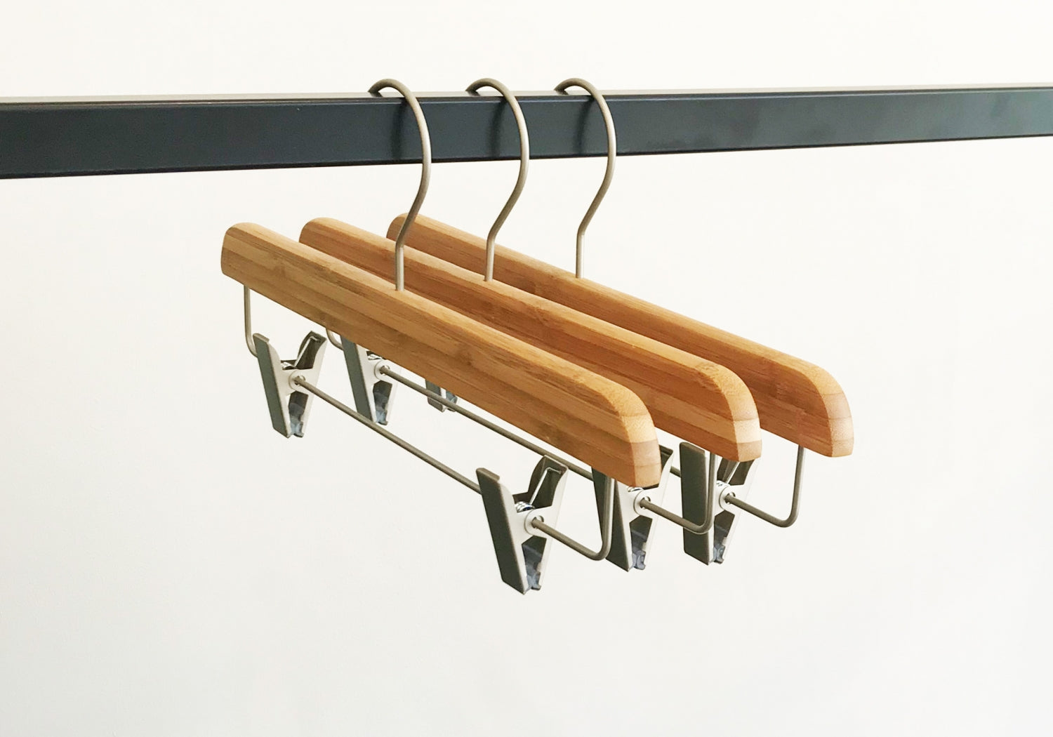 Three Hangers of London Bamboo Wood Sustainable Clip Bottom Hanger 35cm are hanging from a black metal rail. The background is plain white, showcasing these eco-friendly wardrobe accessories.