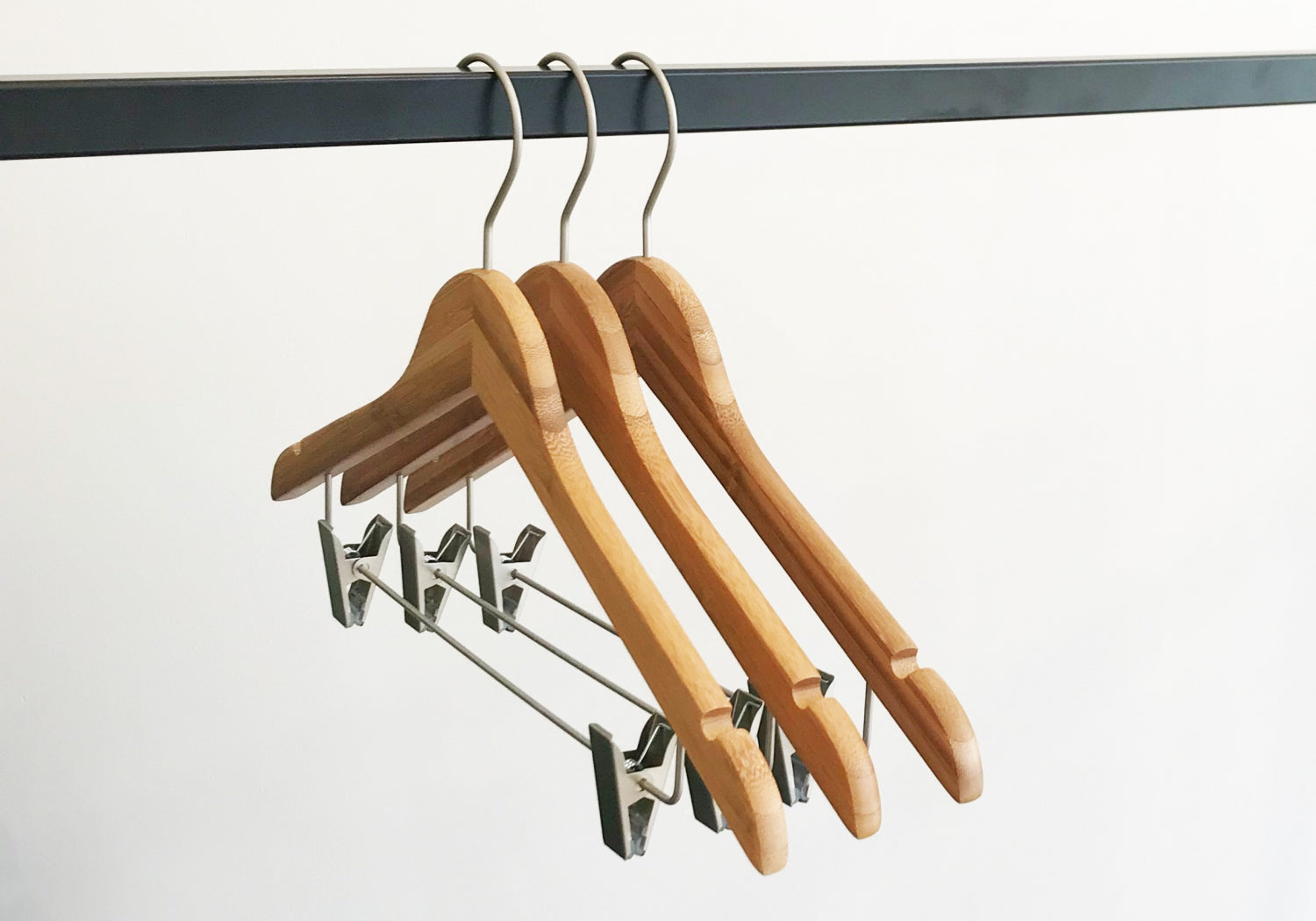 Three Hangers of London Bamboo Wood Sustainable Top Clip Hanger 42cm with metal hooks hang on a black metal rod. The eco-friendly clothing organizers also feature metal clips for securing garments. The background is plain white.