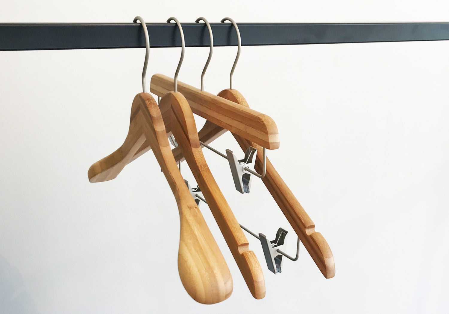 Four Hangers of London Bamboo Wood Sustainable Top Jacket Hanger 42cm with metal hooks hang on a metal rod. Two of the eco-friendly clothing organization tools have curved arms for tops, while the other two feature clips for pants or skirts. The background is plain white.