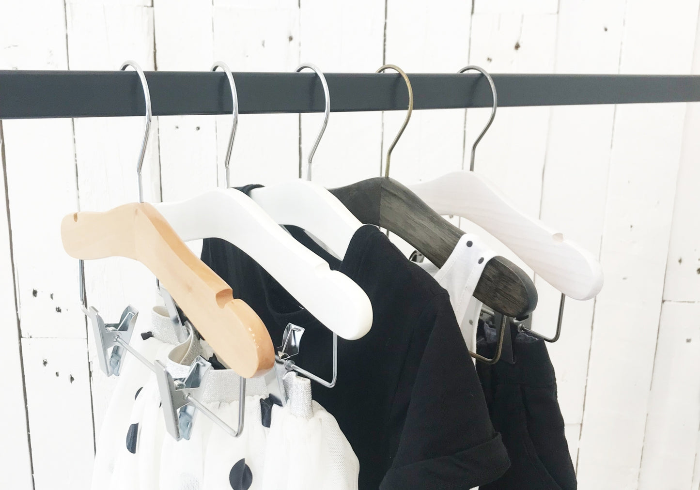 Four 【Hangers of London】 hold a variety of garments on a black rod against a white wooden background. One hanger, complete with metal clips, displays polka dot fabric, while the others showcase black and white clothing items. The 【White Wood Top Coat Kids Hanger with Bar 28cm by Hangers of London】 stands out among the collection.