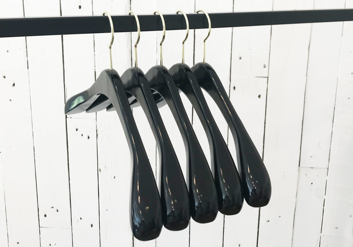 Five high-quality black wood hangers are hanging on a silver metal rod against a white wooden wall. The Hangers of London Black Wooden Top Coat Premium Hanger 42cm are evenly spaced, and the setting appears minimalist. The nonslip grip ensures your clothes stay securely in place.