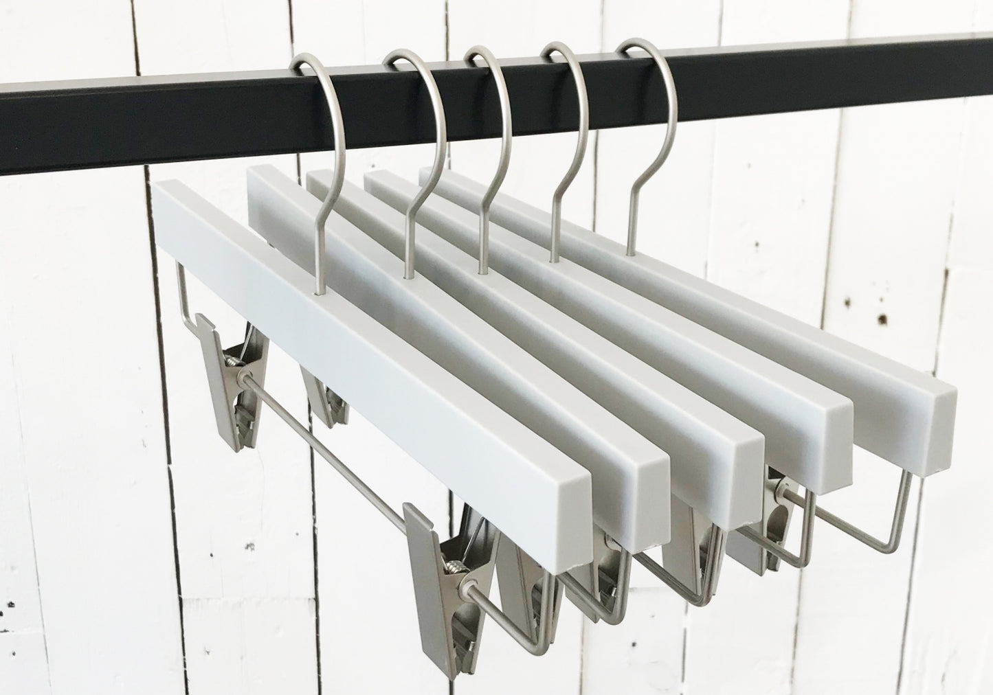 A row of Hangers of London Premium Soft Touch Plastic Clip Hanger 35cm with metal clips and hooks hangs on a black metal rod. The background features a white wooden plank wall, showcasing stylish storage solutions for any wardrobe.
