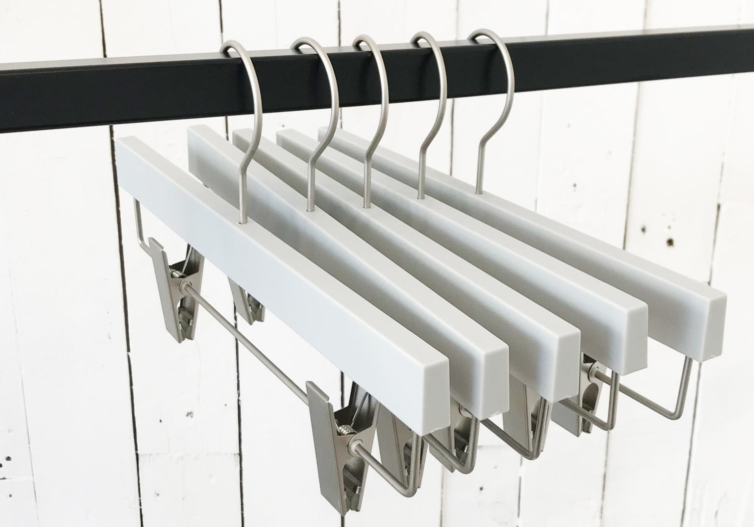 A row of Hangers of London Premium Soft Touch Plastic Clip Hanger 35cm with metal clips and hooks hangs on a black metal rod. The background features a white wooden plank wall, showcasing stylish storage solutions for any wardrobe.