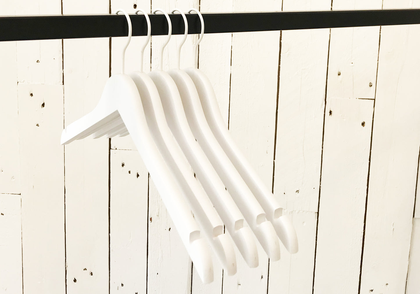 A row of Hangers of London White Wooden Clothes Hanger with Notch 44cm hanging from a black rod against a white wooden panel wall. The high-quality clothes hangers are empty and evenly spaced, creating a minimalist and organized appearance, perfect for closet organization.