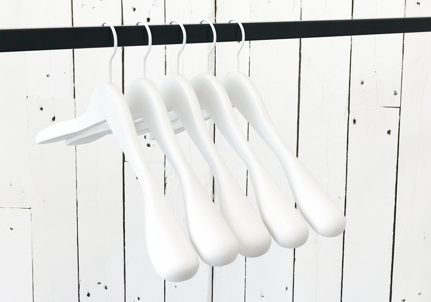 A row of five Hangers of London White Wooden Jacket Top Coat Hanger 44cm hanging on a black rod against a white wooden plank wall. The durable wood hangers are evenly spaced and uniformly aligned, creating a minimalist and orderly appearance.