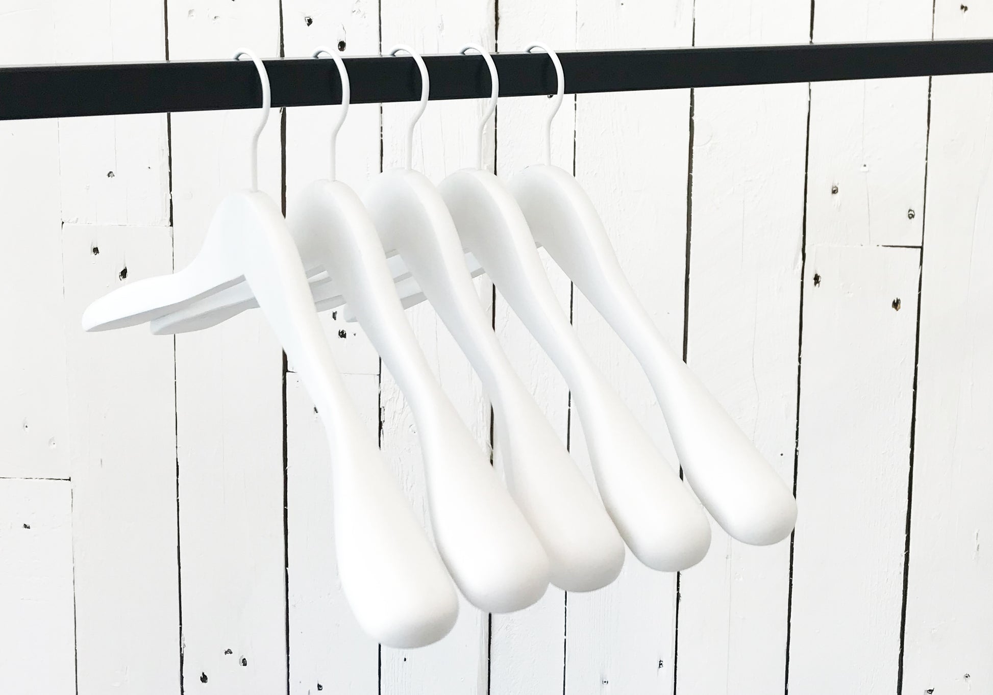 A row of five Hangers of London White Wooden Jacket Top Coat Hanger 44cm hanging on a black rod against a white wooden plank wall. The durable wood hangers are evenly spaced and uniformly aligned, creating a minimalist and orderly appearance.
