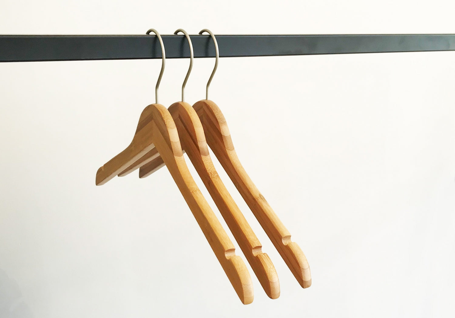 Three Hangers of London Bamboo Wood Sustainable Top Hangers with Notch 42cm hang on a black metal rail against a plain white background. The hangers are evenly spaced and aligned, with their metal hooks visible at the top, creating a simple and minimalist composition.