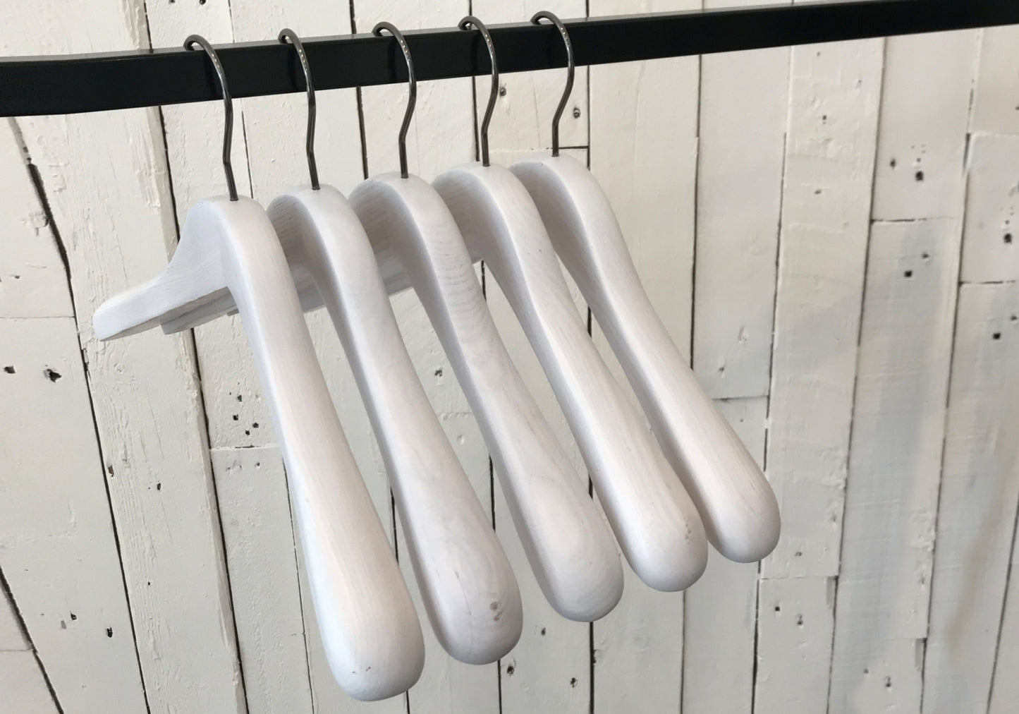 Five premium-quality White Ash Wooden Jacket Coat Hanger 44cm by Hangers of London are hanging on a black metal rod against a background of white wooden planks.