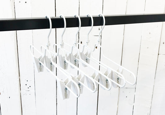 Five stylish White Metal Clip Bottom Hanger 35cm from Hangers of London are hanging from a metal rod. The background consists of white wooden panels, giving a minimalist and clean appearance.